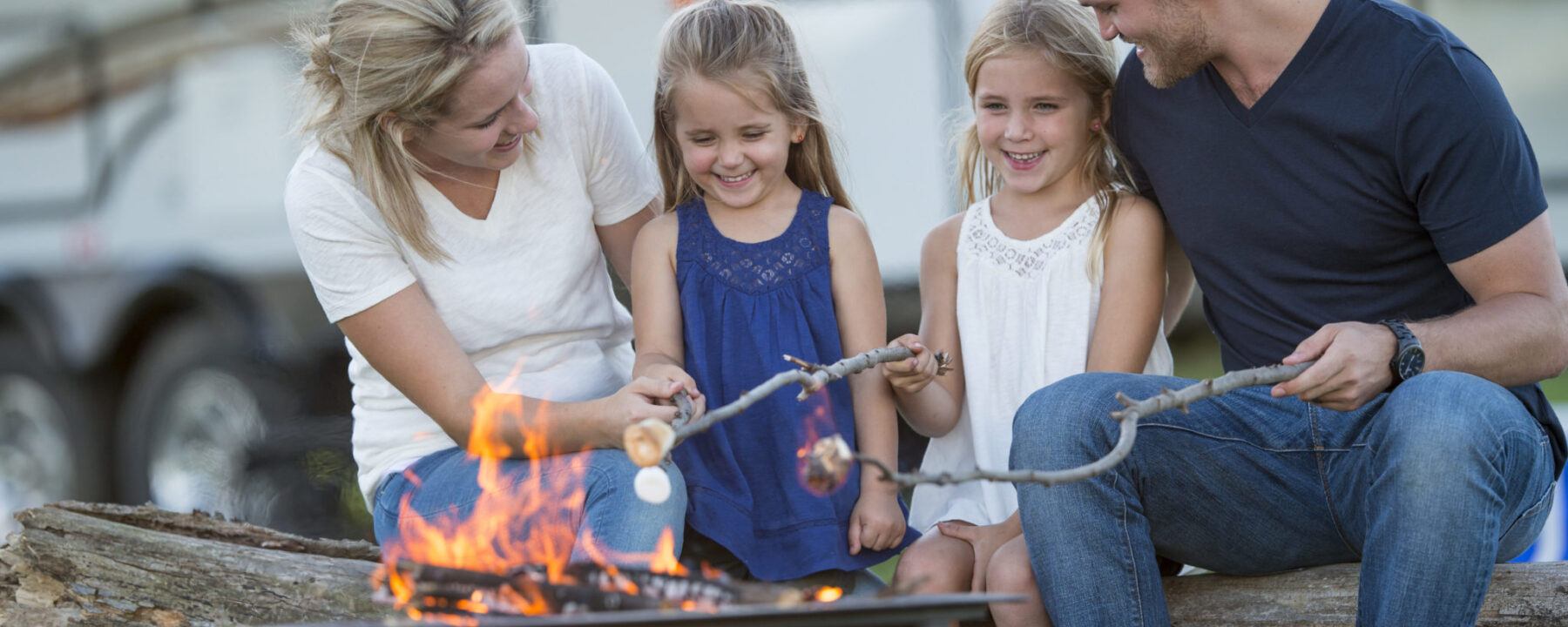 A family of four are roasting marshmallows together around a campfire while on vacation. Their RV is parcked in the background of the picture.